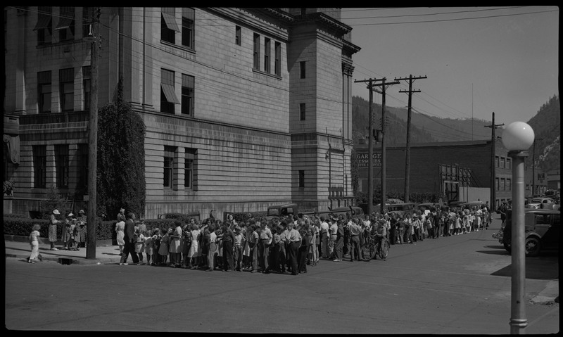Many children gather in the street near a large building for a back-to-school parade. Some adults are standing on the sidewalk.