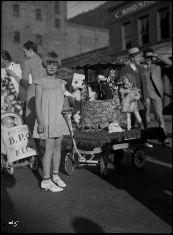 A child poses with her decorated wagon during the Benevolent and Protective Order of Elks parade. Her wagon is decorated with a well, a doll, and a fake rabbit. Other parade participants and spectators are standing in the background. 