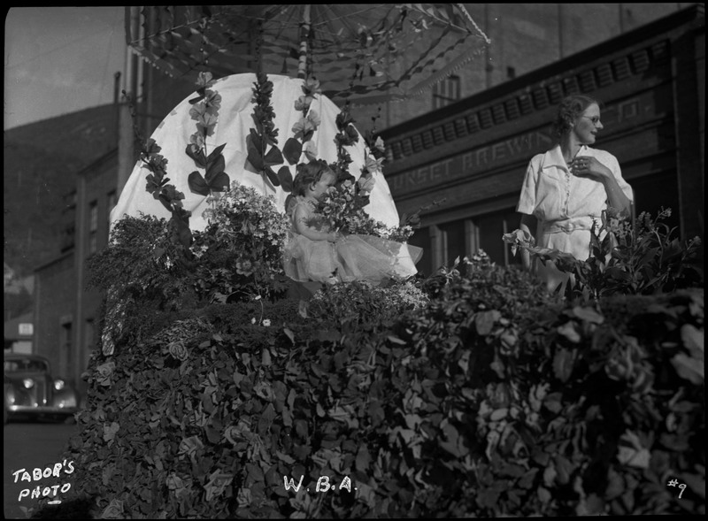 A girl and a woman on the W.B.A. float during the Benevolent and Protective Order of Elks parade. The float is decorated in foliage.