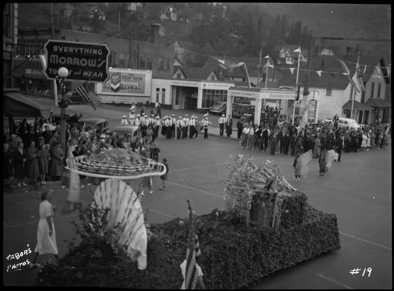 Street view of a line of decorated floats and parade participants in costume during the Benevolent and Protective Order of Elks parade. Crowds of spectators watch the parade from the sides of the street.