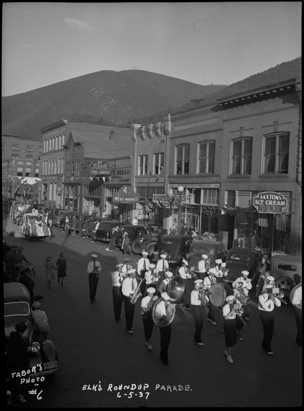 Marching band performing during the Benevolent and Protective Order of Elks roundup parade. Floats follow the marching band as spectators watch from the sidewalk and near parked cars.