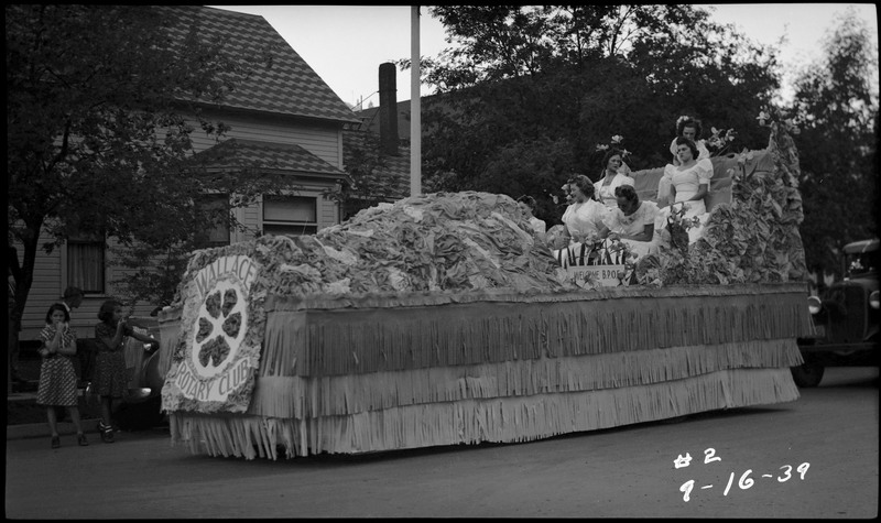 Six women sitting in the Wallace Rotary Club float during the Benevolent and Protective Order of Elks roundup parade. Two girls can be seen watching the parade on the right, a boy stands behind them facing away.