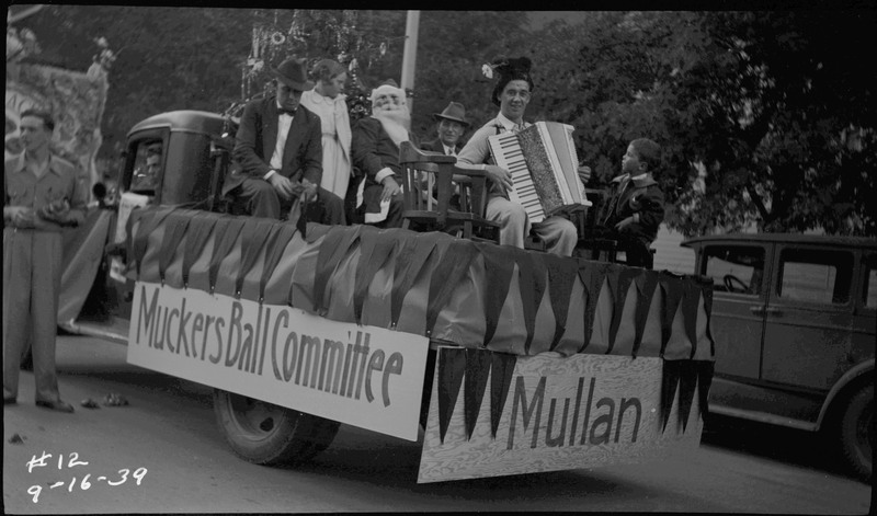 Six men and children sitting on the Muckers Ball Committee float. The sign on the back of the float reads, "Mullan." One of the men is dressed up as Santa Claus and another man holds an accordion.