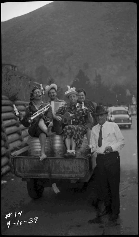 Four men, dressed up as women, sitting on barrels on a flat during the Benevolent and Protective Order of Elks parade. Another man stands near the vehicle.