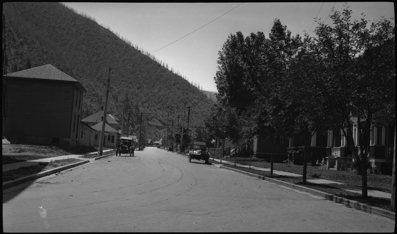 View of an empty street during the Benevolent and Protective Order day parade, There are a few automobiles parked on the street. Trees and building are in view.