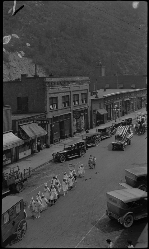 A line of children wearing white capes, dresses, and hats follow an ambulance during the Benevolent and Protective Order of Elks day parade. One child is holding a sign that reads "Health Crusade." Two other vehicles follow the children and spectators watch from parked cars on the street and the sidewalk.