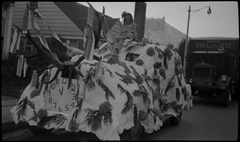 A girl sitting on the top of a automobile decorated as a float with tree branches and fake antlers during the Benevolent and Protective Order of Elks day parade.