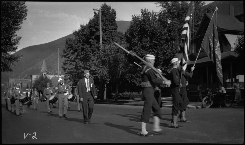 People in uniforms marching during the V.F.W. State convention parade, some are holding flags and rifles on the right. Other uniformed people are holding instruments in the background. 