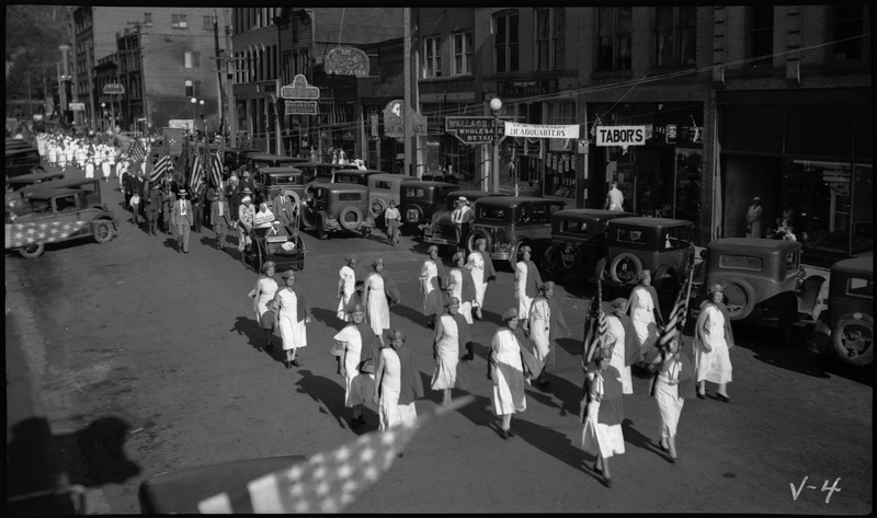 Women in matching uniforms with capes during the V.F.W. State convention parade. Other people, including a man in a wheelchair, follow the women in the parade procession. Spectators watch from the sidewalk.  
