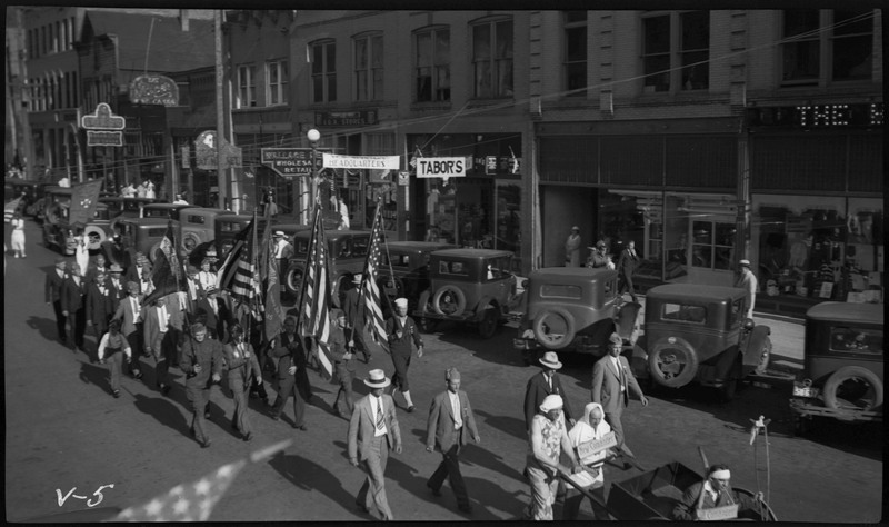 Men in uniforms, six holding flags during the V.F.W. State convention parade. Two people are wheeling a man sitting in the wheelchair on the right. Spectators can be seen watching from the sidewalk. Several storefronts, including Tabor's are in the background.