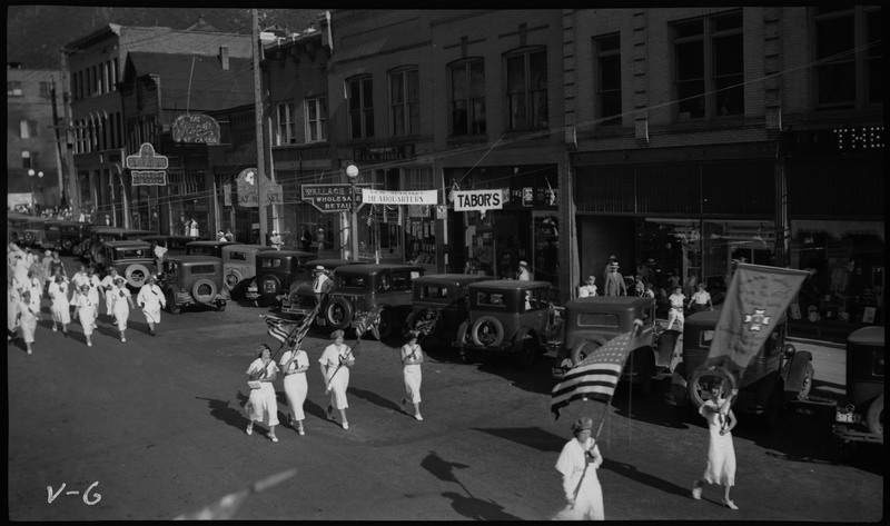 Women wearing white, six holding flags, walking in the V.F.W. State convention parade. Several storefronts can be seen in the background, including Tabor's. Cars are parked along the side of the street.