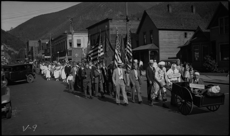 A crowd including men in uniform, women wearing white, and a man in a wheelchair during the V.F.W. State convention parade.