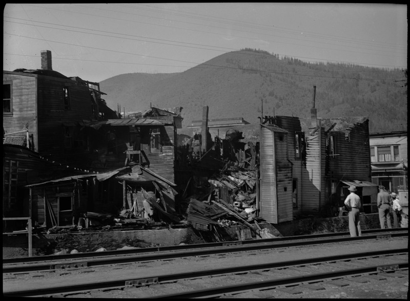 A rear view of damaged buildings on the east side of 6th street after a fire. Three people can be seen surveying the damage on the right.