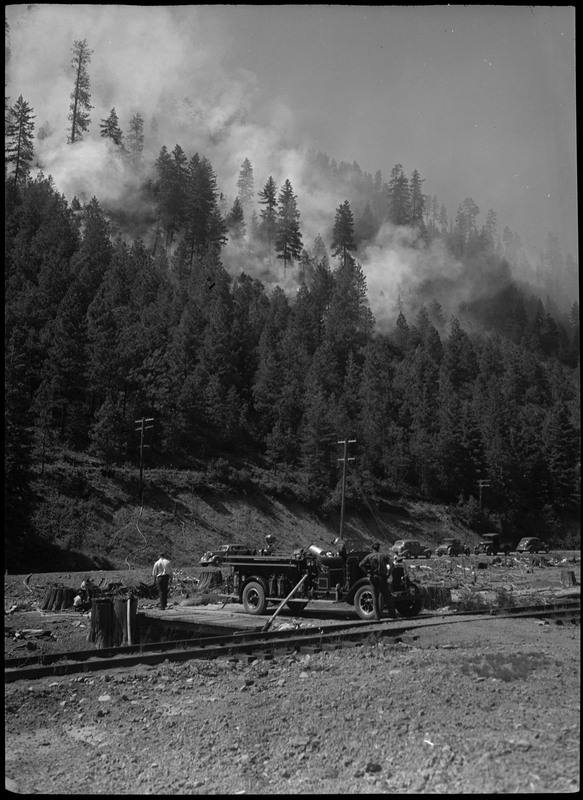 A group of people stand near a fire truck and look on at the smoke rising from a forest fire outside Wallace. A few other cars are visible on a nearby road.