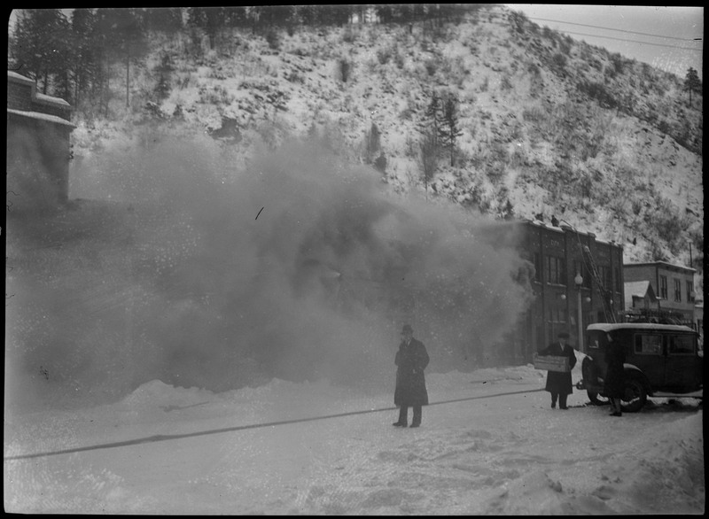 Smoke almost entirely obscures the McKinley Building while it's on fire. Two people are visible in the foreground. One of them is carrying a box, the other appears to be smoking a pipe. Firefighters can be seen on a nearby building, and a firetruck is on the street.