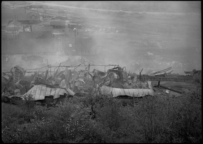 Remnants of a building including metal and broken machinery after a fire at Morning Mill. Smoke clouds part of the view. Some standing buildings can be seen in the distance and vegetation in the foreground.