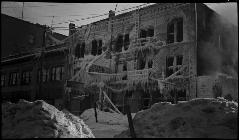 Soot and what appear to be icicles and frost cling to the structure of a building after a fire. There appear to be no window panes anymore, and there are piles of snow and soot on the ground.