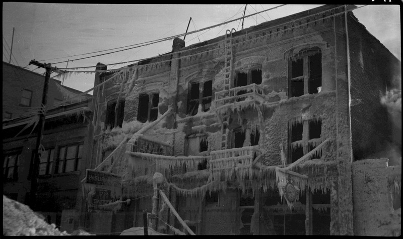 View of the Ryan Hotel and Tabor Building after a fire. Soot and what appear to be icicles and frost cling to the structure of a building after a fire. There appear to be no window panes anymore, and there are piles of snow and soot on the ground.