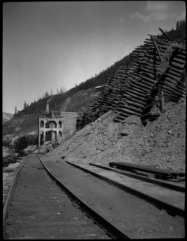 View of an empty railroad track with a structure at the end in the distance, situated by the side of a hill. Stacks of logs are sticking out from the hillside.