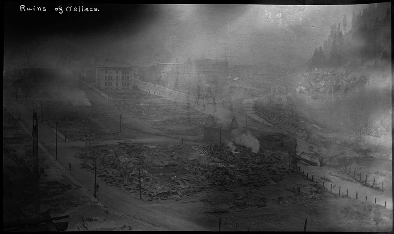 Ruins of Wallace after a fire in 1910. A smoky haze hangs over the ruins of buildings, while some buildings appear to still be standing.