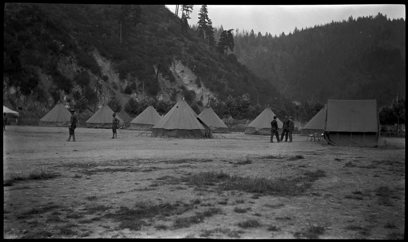 Five men standing near tents with a forest landscape in the background, possibly around the time of the 1910 Wallace fire.