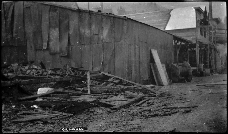 Cil House after the 1910 Wallace fire. There are ruins next to a standing building made of corrugated metal.
