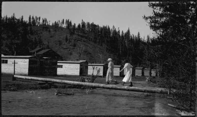 Two people walk across a log over a body of water, possibly around the time of the 1910 Wallace fire.