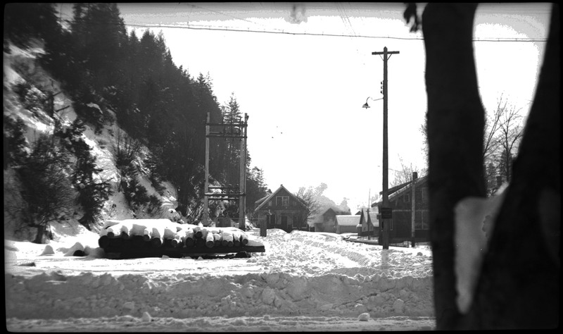 Snowy street scene around the time of the Pearson house fire. There are houses and a pile of logs in view.