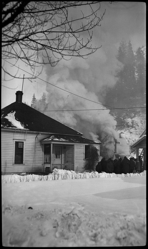 People gather near the Pearson house fire, where heavy smoke can be seen rising from the house. There is also snow in the foreground.