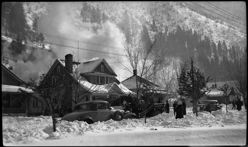People walking near or watching the Pearson house fire from the street. There are cars parked along the snowy street and a snow covered hill in the background.