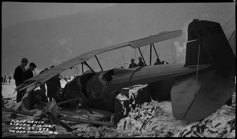 Several people look at and stand near the remnants of an airliner crash in the snow. The tail numbers are "C 6156." Caption on the bottom left of the photograph reads "Plane crash Osburn Airport Dec. 27, 1933 Tabor Photo A6."
