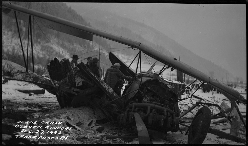 Three unidentified men and one woman (with a dog) inspect the remnants of an airliner crash in the snow. Caption on the bottom left of the photograph reads "Plane crash Osburn Airport Dec. 27, 1933 Tabor Photo A5."