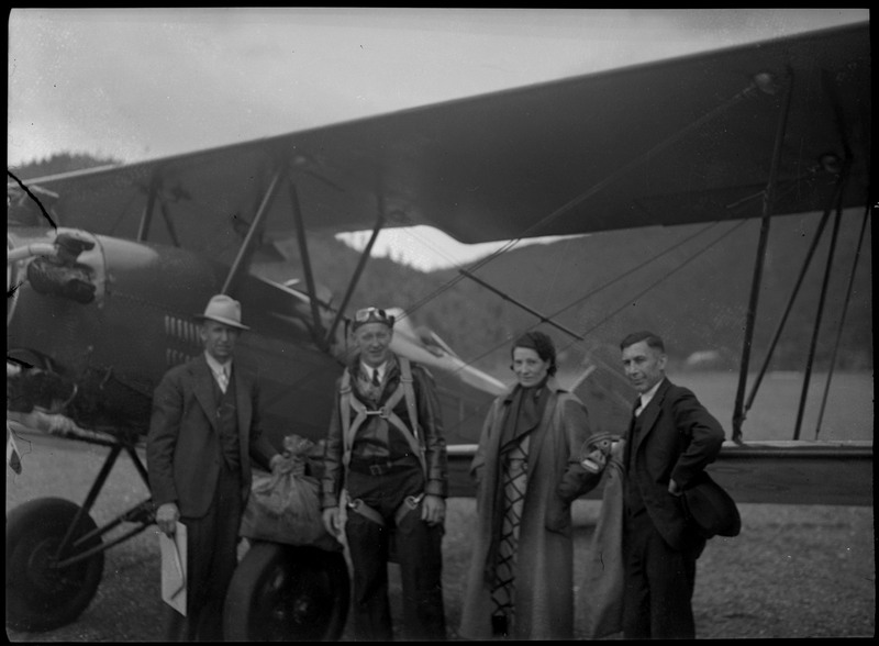 Pilot, George Nelson, stands in front of an airplane with one woman and two men. One holds a bag and large envelope.