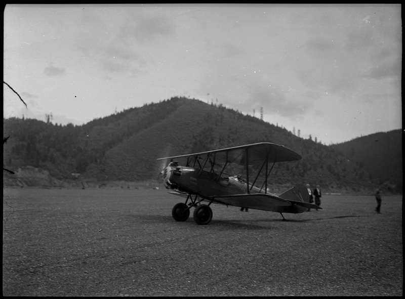 An airplane in an empty field with trees and mountains in the background. Three unidentified people stand behind it.