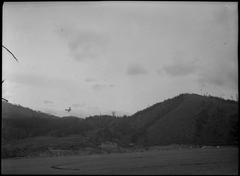 View of airplane (on the left) over a forested landscape as it takes off for its first mail flight.