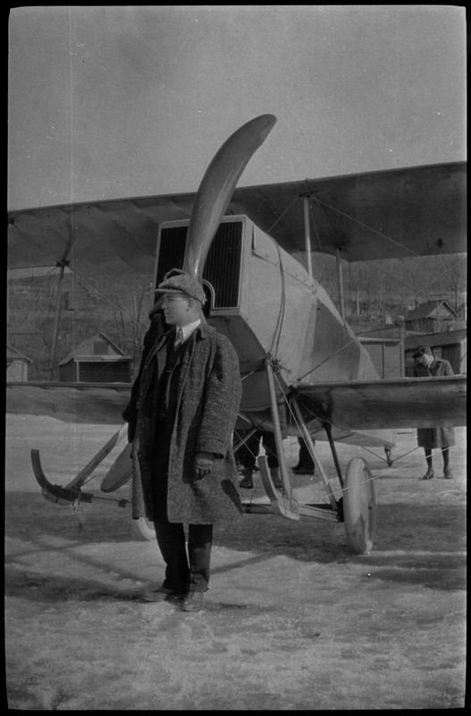 A man wearing a hat and coat stands in front of the FLA airplane. Other people can be standing behind the plane.