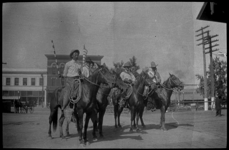 Four men sitting on horses in the street with buildings in the background.  There is possibly a fifth person on a horse right behind them.