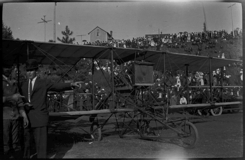 Two men stand on the left in front of an airplane. Many people are gathered on a hillside in the background.