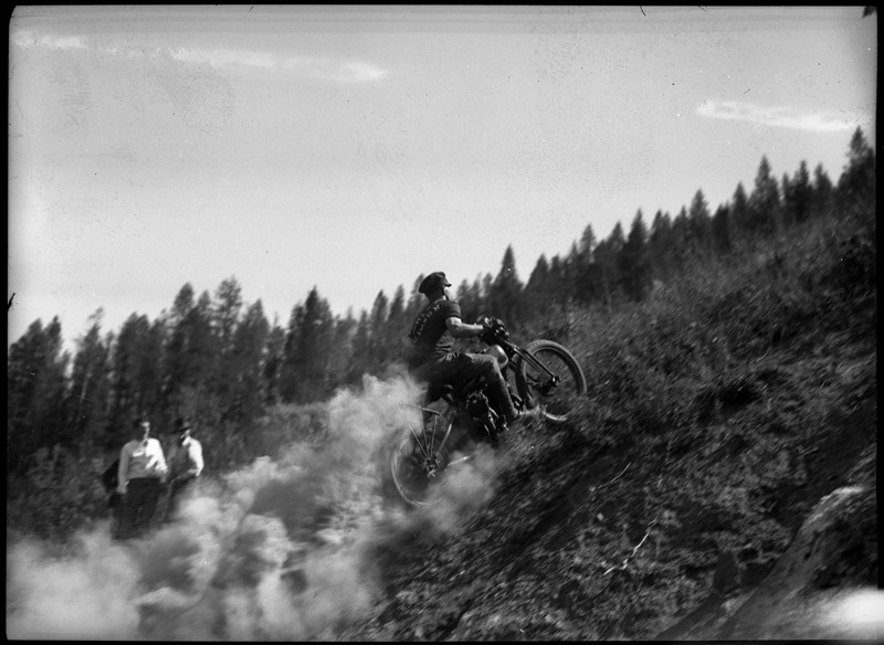 A man on motorcycle hill climb. Dust trails the motorcycle as two others watch on the left.