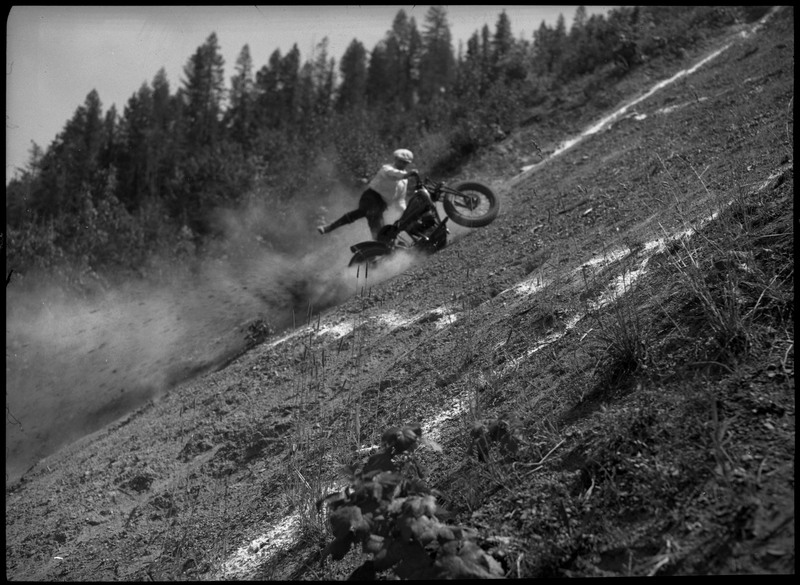 A blurry image of a man is on a tilted motorcycle as others watch along the slope of the hill. There are white markings along the hill.