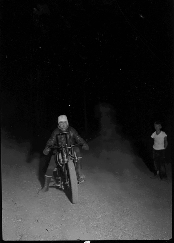 A man wearing a helmet faces the camera on a motorcycle during a hill climb. A boy watches on the right. They are both surrounded by darkness.