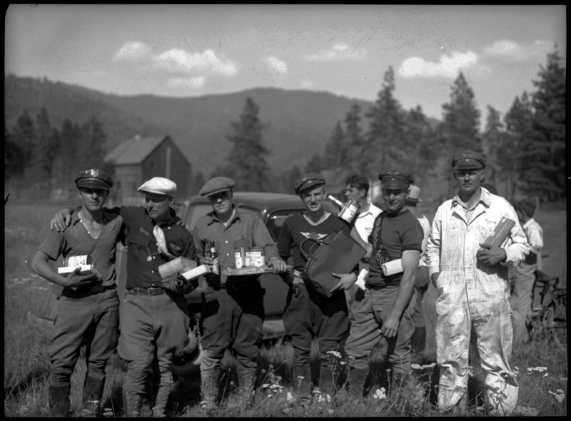 Six men pose while holding food and other things in their hands. There is a car and other people standing behind them. This image was taken during a motorcycle hill climb.
