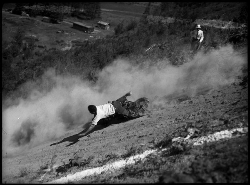 A man falling off his motorcycle during a hill climb. There are markings along the hill. Dust and dirt is flying in the air from the motorcycle. A man can be seen watching on the right.