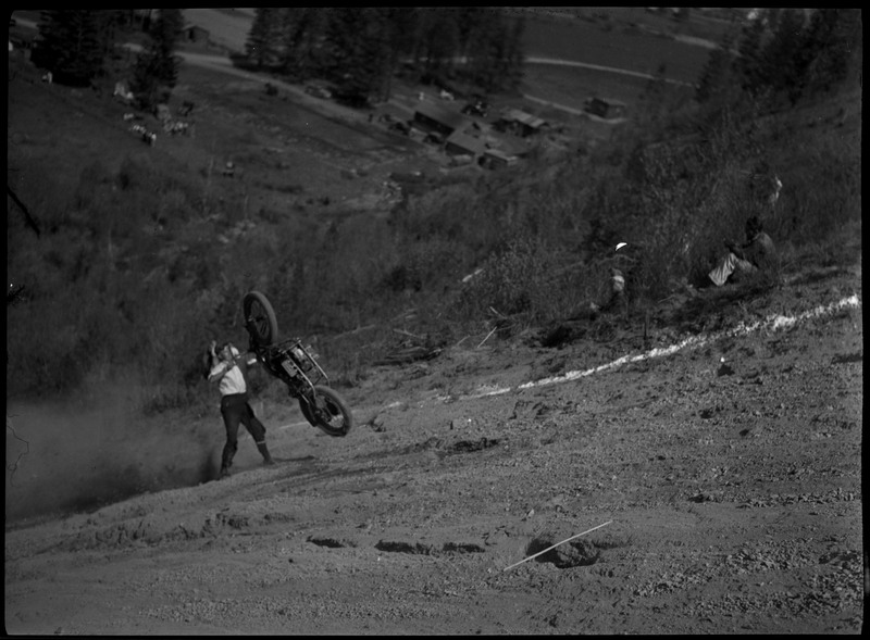 Man falls backwards from a motorcycle after attempting a hill climb. Dust trails behind him. People can be seen watching from the sides.