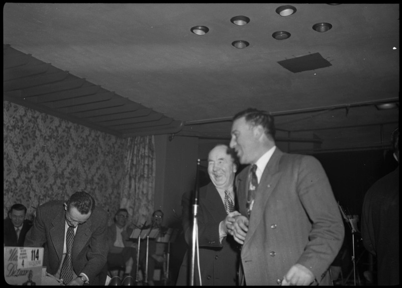 Two men standing and laughing at the March of Dimes event held at the B.P.O.E.temple. There are band members in the background, seated.