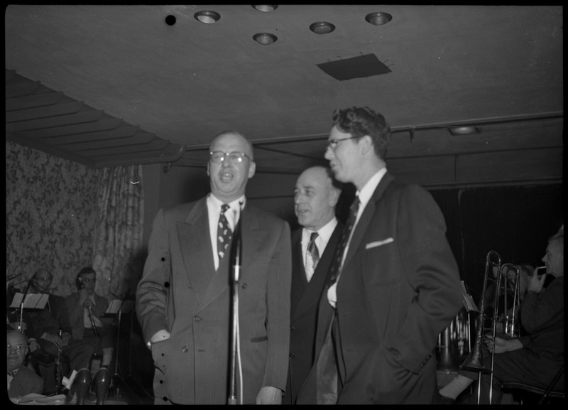 Three men standing around a microphone at a March of Dimes event held at the B.P.O.E. temple. Members of a band can be seen seated with their musical instruments in the background.