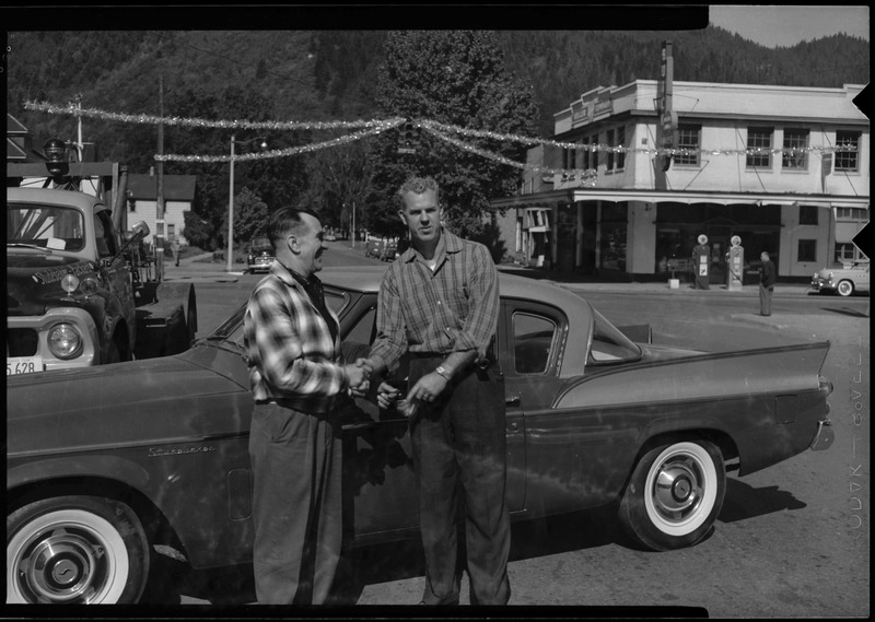 Two men shaking hands in front of a car. A gasoline pump can be seen in the background.