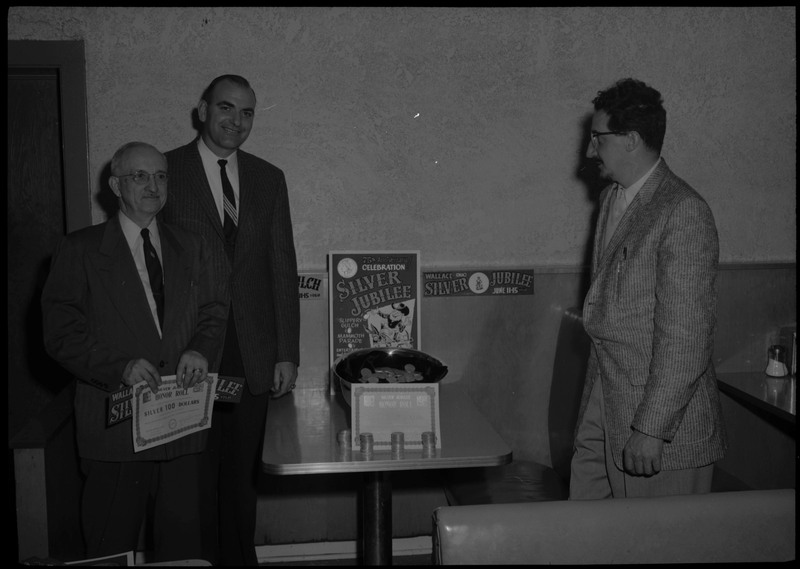 Three men standing around a Silver Jubilee display during the Silver Jubilee kickoff banquet.