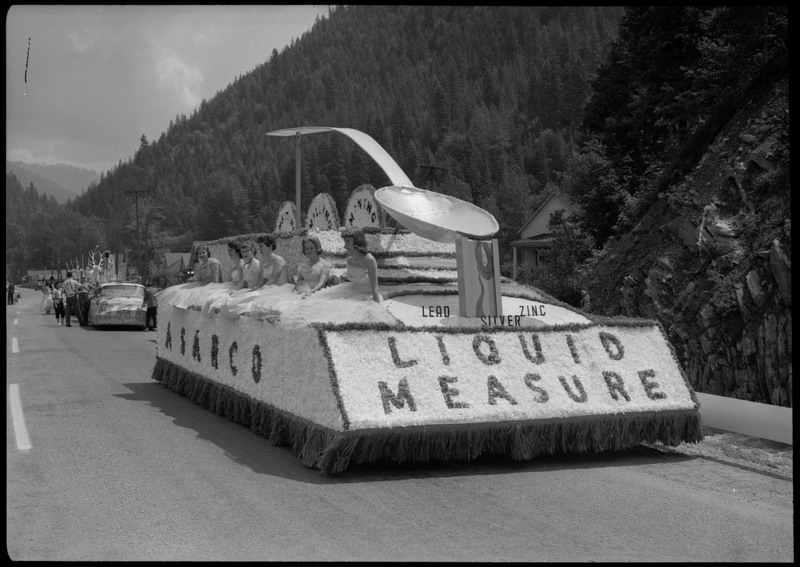 Six women dressed in gowns sitting on an ASARCO float during the Silver Jubilee. The float is decorated with a giant replica of a spoon. Text along the float reads, "ASARCO," "Lead Silver Zinc" and "Liquid Measure." 