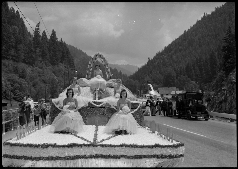 Five women dressed in gowns sitting on a float during the Silver Jubilee. Bystanders and parade participants are nearby.